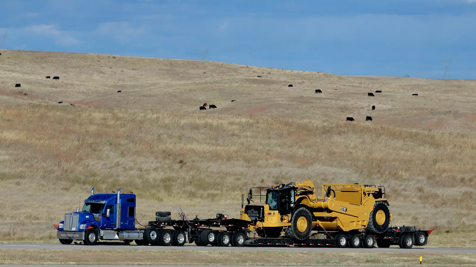 Truck pulling a multi-axle low-bed trailer carrying heavy earthmoving equipment along a rural road