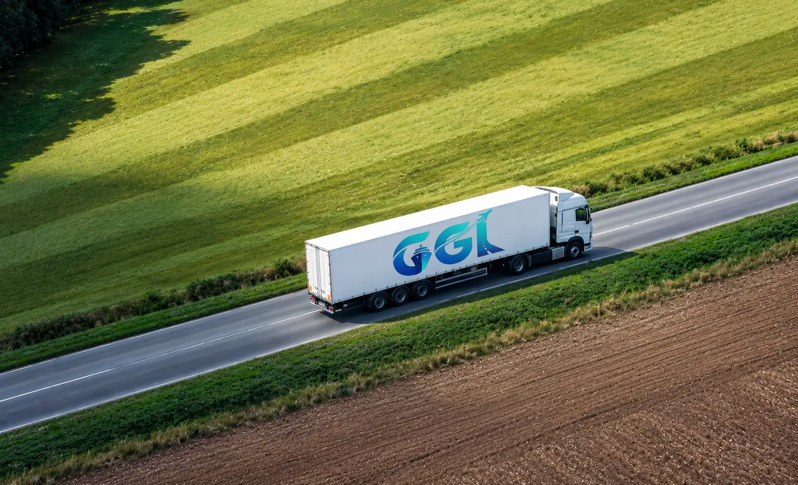 Truck with a GGL-branded trailer on a straight road between green and ploughed fields, seen from the air