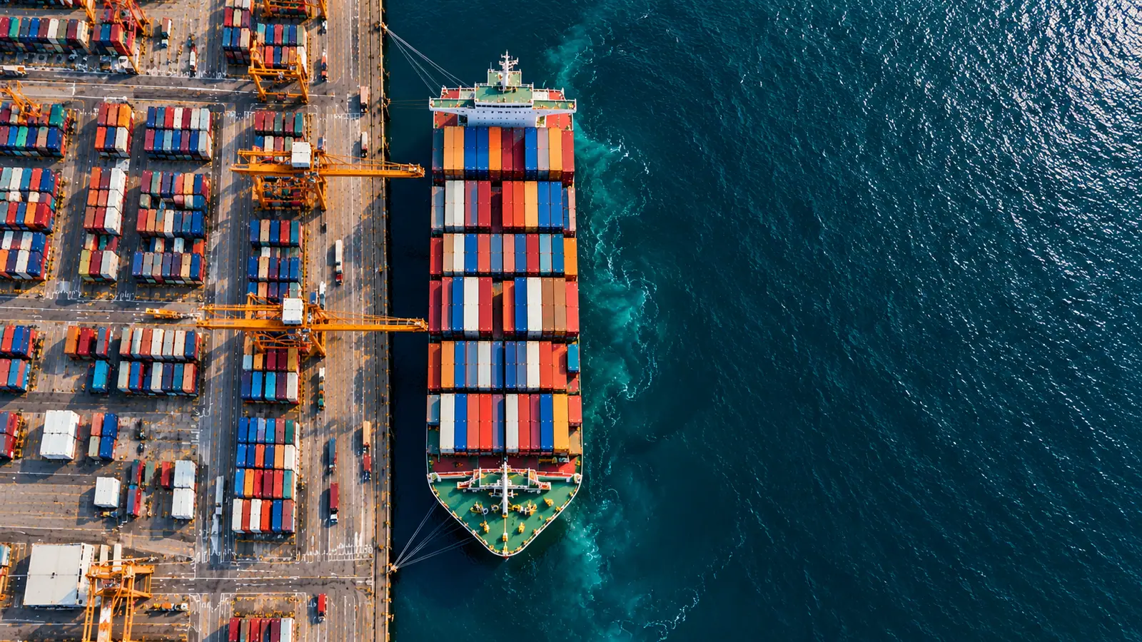 Container ship berthed at a terminal beside container stacks and gantry cranes, seen from above
