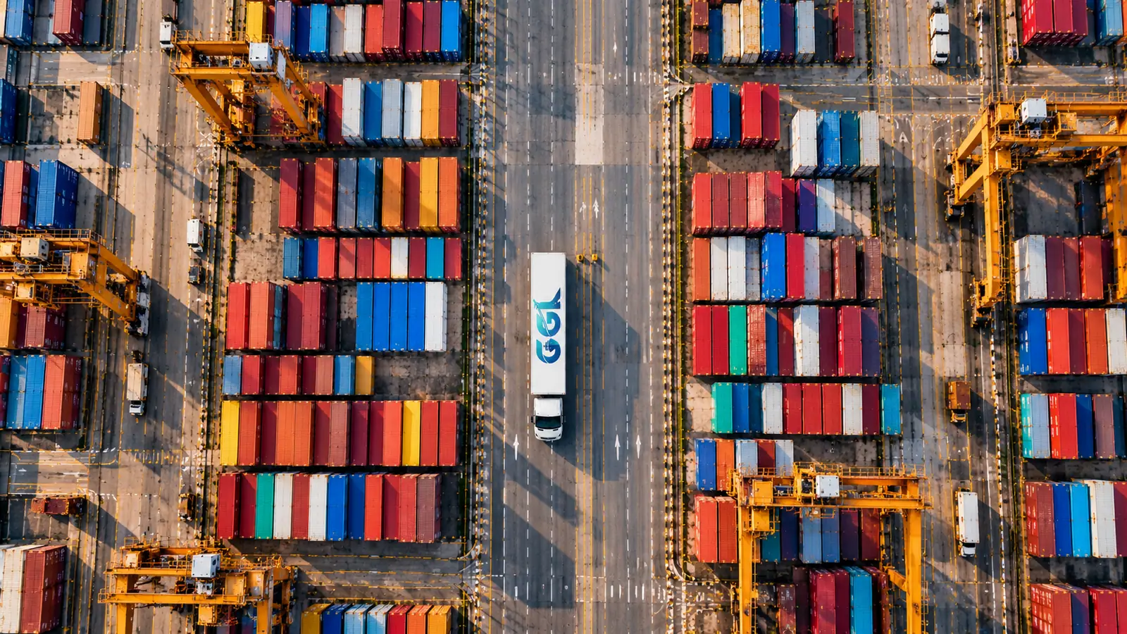 Truck with a GGL-branded trailer driving down the road between container stacks and gantry cranes, seen from above